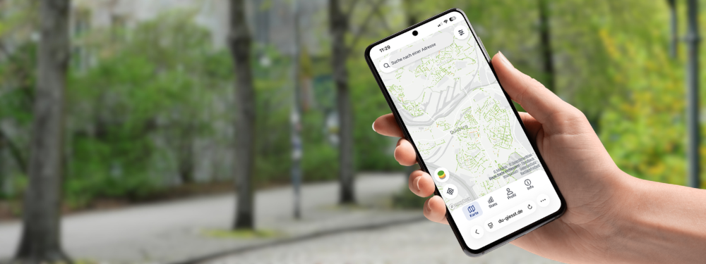 Eine Hand hält ein Handy mit der Ansicht von DU giesst, im Hintergrund Straßenbäume.