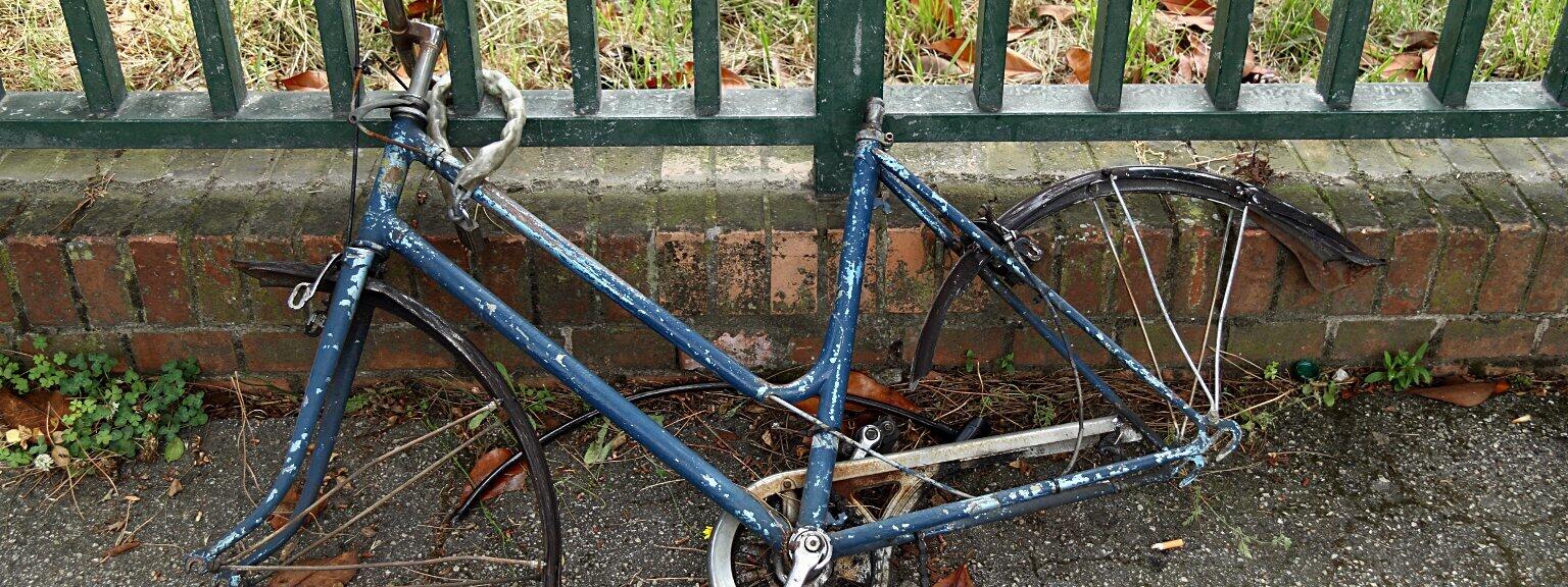 Ein kaputtes Fahrrad ohne Reifen an einen Zaun angekettet.