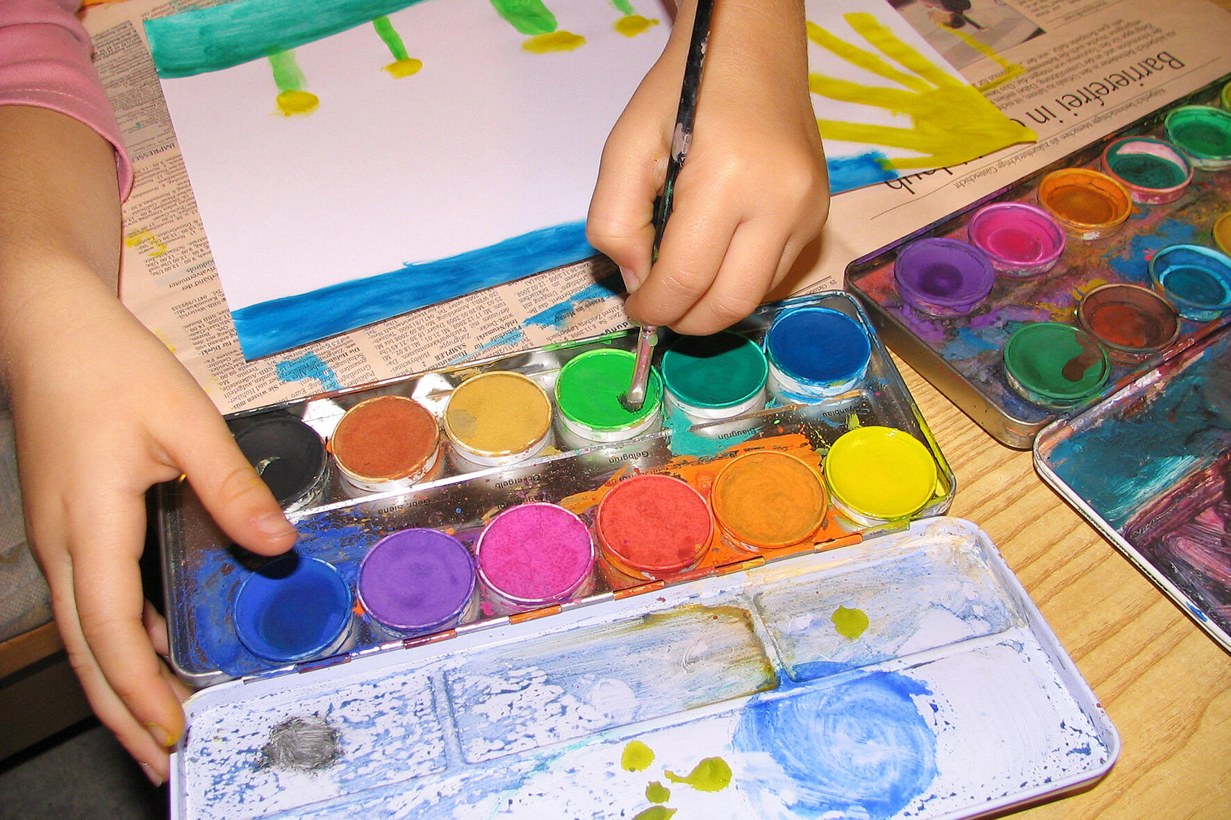 Ein Kind malt ein Bild. Zu sehen sind die Hände und bunte Tuschkästen mit Wassermalfarbe