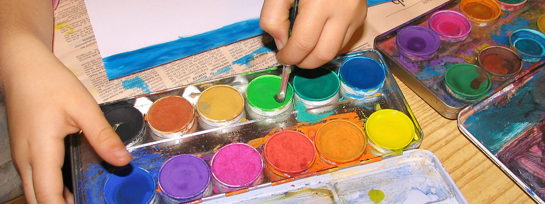 Ein Kind malt ein Bild. Zu sehen sind die Hände und bunte Tuschkästen mit Wassermalfarbe