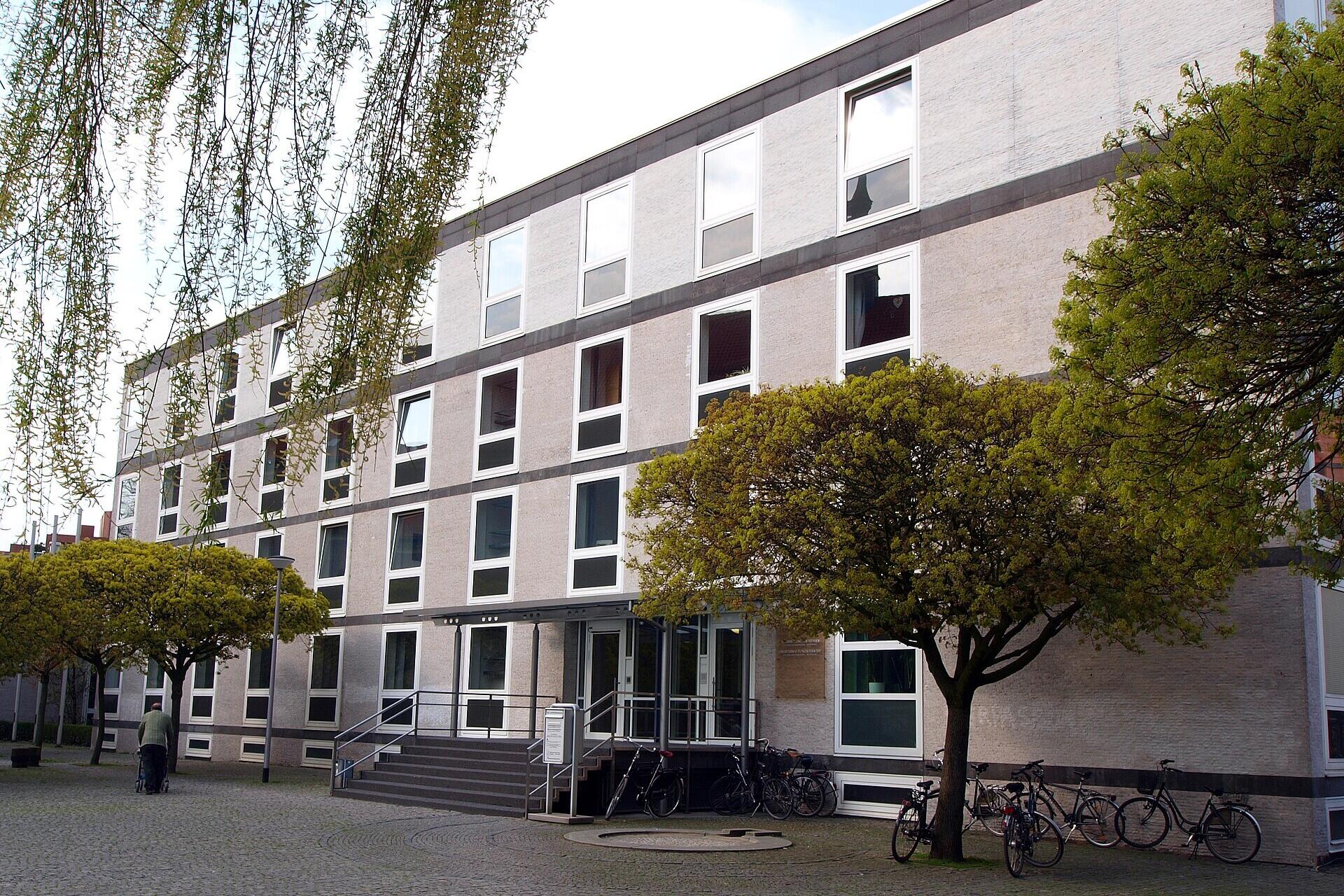 Gebäudeansicht OVG Münster. Nüchterne und schmucklose Fassade, im Vordergrund mit Bäumen und Fahrrädern