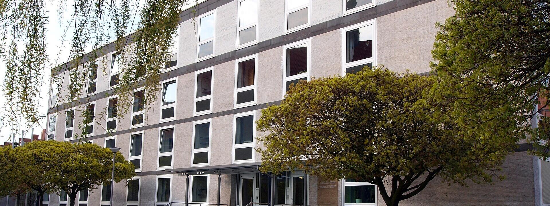 Gebäudeansicht OVG Münster. Nüchterne und schmucklose Fassade, im Vordergrund mit Bäumen und Fahrrädern