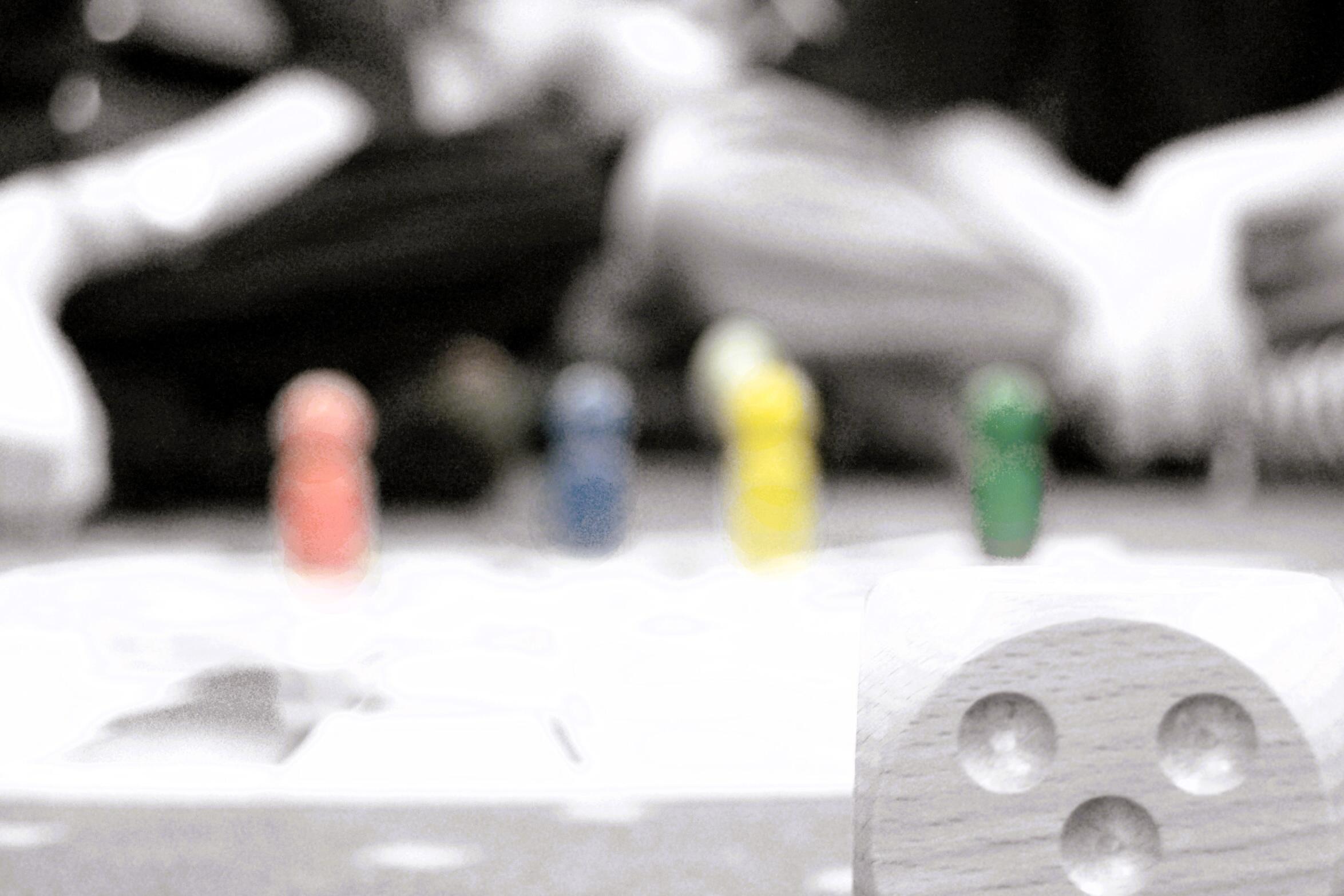 Ein Würfel im vordergrund, Spielfiguren und sitzende Personen im Hintergrund