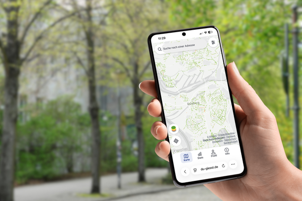 Eine Hand hält ein Handy mit der Ansicht von DU giesst, im Hintergrund Straßenbäume